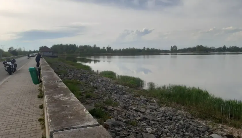 Nad bralinem, gdzie woda odbija chmury, stoi człowiek. Obok motocykle i ścieżka z kostki brukowej.