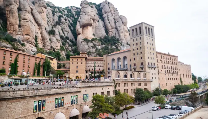 Edificios del monasterio se alzan bajo las imponentes rocas del parc natural de la muntanya de Montserrat. Turistas pasean por la plaza.