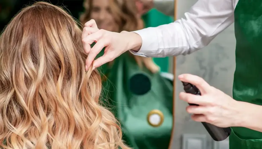 Fryzjerka utrwala fryzurę klientki, spryskując jej blond włosy lakierem. Perfekcyjny styling włosów.
