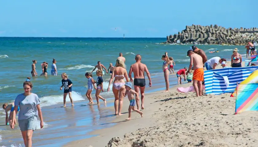 Rodziny cieszą się słońcem i morzem. Widać plażowiczów, dzieci bawiące się w wodzie. Ceny nad morzem w wakacje są wysokie, ale warto.