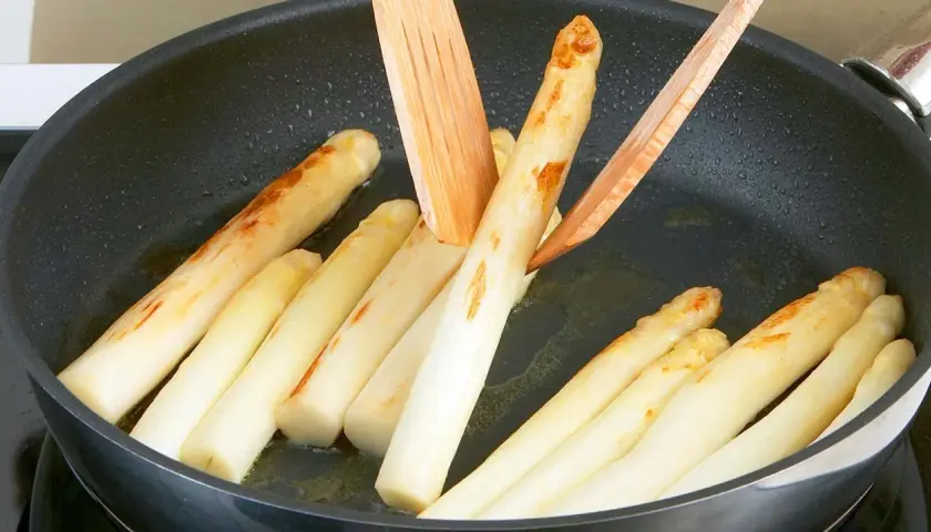 Weißer Spargel brät in einer Pfanne mit etwas Öl. Holzspieße wenden die Stangen, die leicht gebräunte Stellen zeigen.