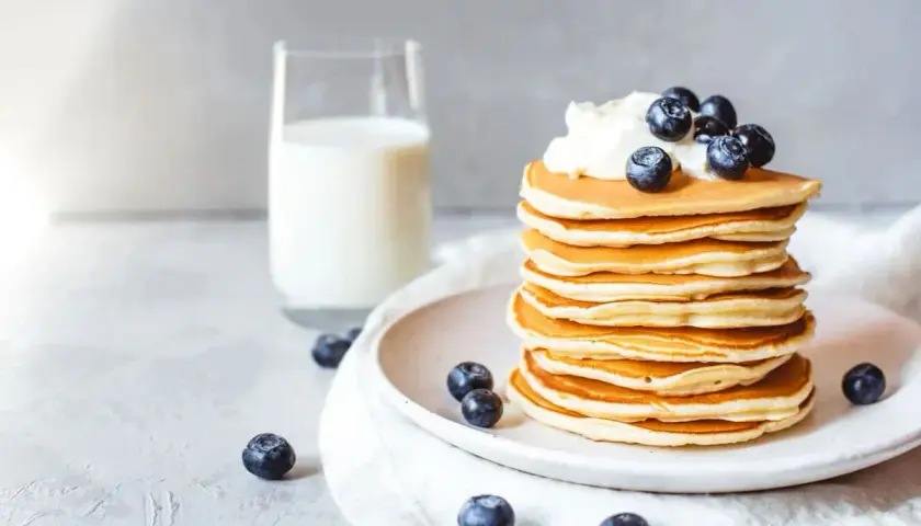 Ein Stapel goldbrauner Protein Pfannkuchen, garniert mit Sahne und Blaubeeren, daneben ein Glas Milch.