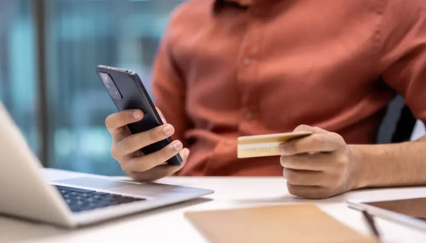 Osoba trzymająca telefon i kartę kredytową przy laptopie.