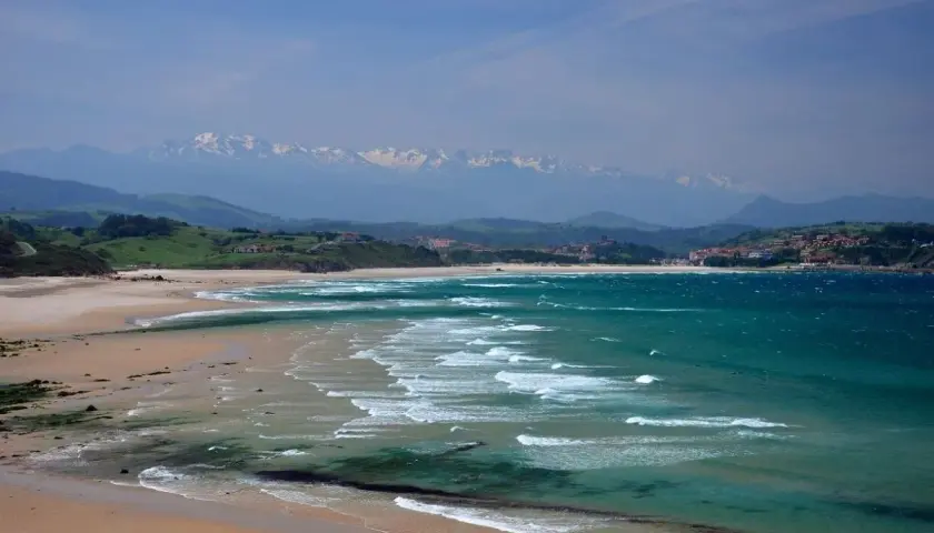 Amplias playas de arena dorada y aguas turquesas, con olas rompiendo suavemente. Al fondo, colinas verdes y montañas nevadas, un paisaje idílico de playas cerca de Comillas.