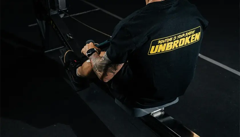 Mężczyzna z tatuażami na ramieniu trenuje na wioślarzu. Czy to crossfit czy siłownia, ważne, że jest UNBROKEN!
