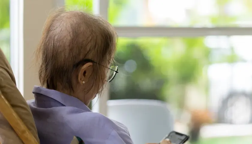 Osoba po chemioterapii w okularach siedzi przy oknie, trzymając telefon.