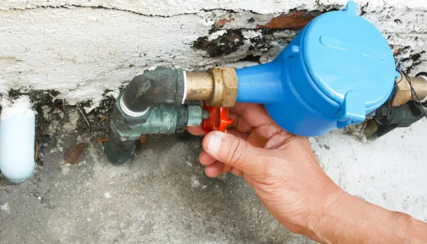 Main d'une personne actionnant le robinet rouge du tuyau d'arrivée d'eau de la maison, juste avant le compteur bleu.