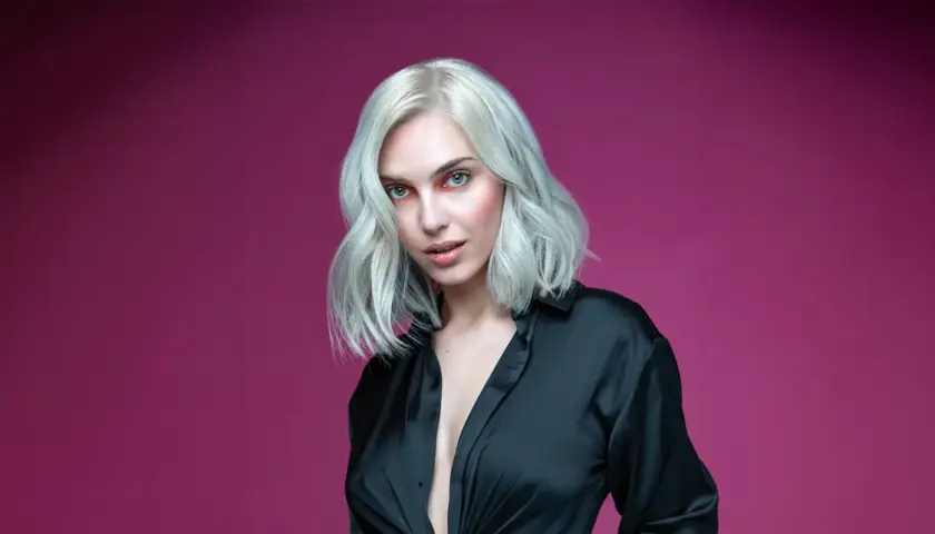 Mujer con cabello rubio ceniza platino, ojos azules y camisa negra abierta, sobre fondo rosa.