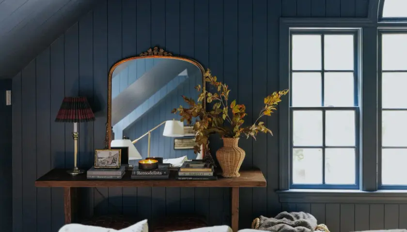 A rustic style console table with a lamp, books, and a vase of autumn leaves sits against a dark blue paneled wall, reflecting in an ornate mirror.