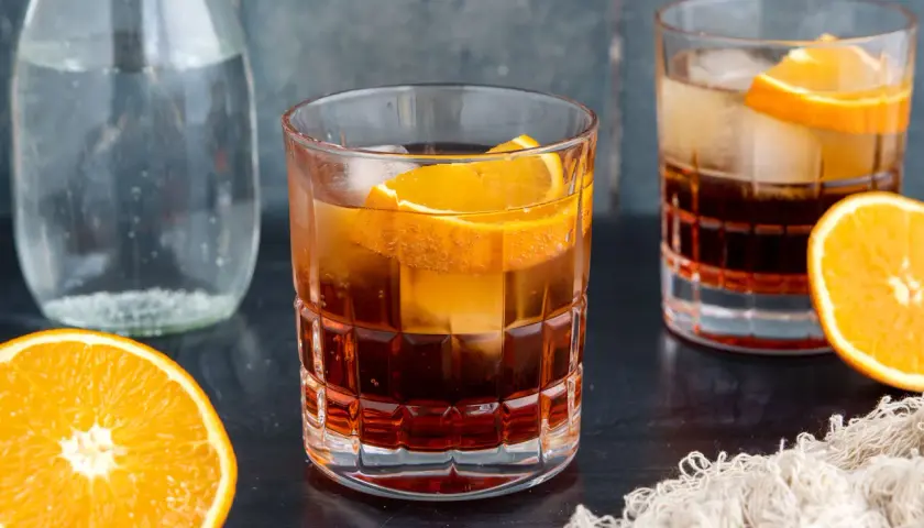Ein erfrischender Americano Cocktail mit Eis und Orangenscheiben, daneben eine Karaffe Wasser und frische Orangen.