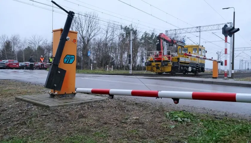 Żółta lokomotywa z dźwigiem zbliża się do przejazdu kolejowego bez zapór. Na pierwszym planie pomarańczowy słup z podniesionym ramieniem.