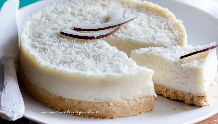 Delizioso cheesecake al cocco, ricoperto di cocco grattugiato e decorato con scaglie di cocco fresco. Una fetta è già stata tagliata, invitando a gustare questo dolce tropicale.
