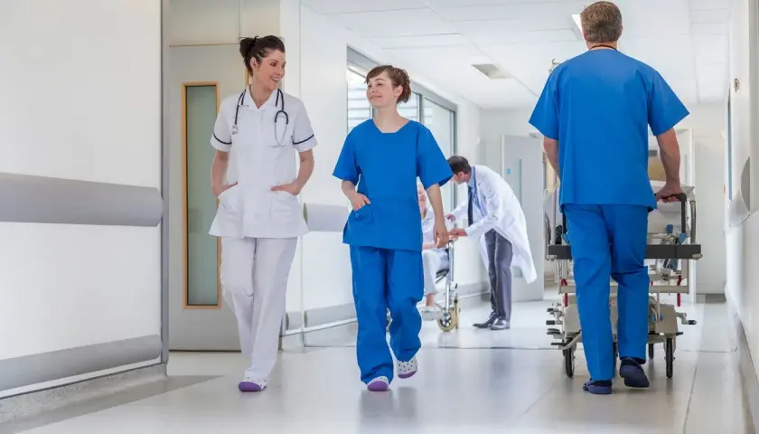 In un corridoio ospedaliero, personale medico si muove con urgenza. Un uomo spinge una barella, mentre due infermiere camminano velocemente, forse a seguito di un evento avverso.