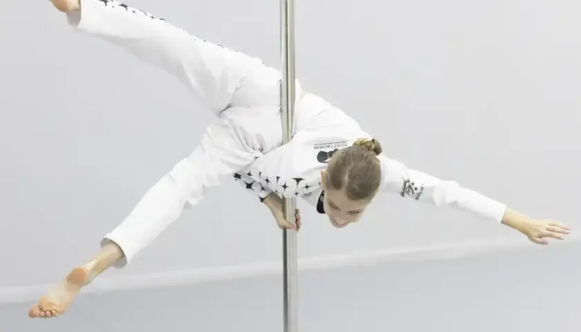 Dziewczyna w białym stroju z gwiazdami wykonuje akrobatyczną figurę na rurze. Czy pole dance to grzech? Ta młoda sportsmenka udowadnia, że to sztuka.