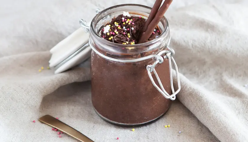 Un pot de mousse au chocolat mascarpone, garni de vermicelles colorés et de bâtonnets au chocolat. Une cuillère dorée repose à côté.
