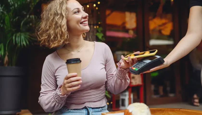 Uśmiechnięta kobieta z kawą płaci telefonem zbliżeniowo za croissant. Szybki przelew na telefon w kawiarni.