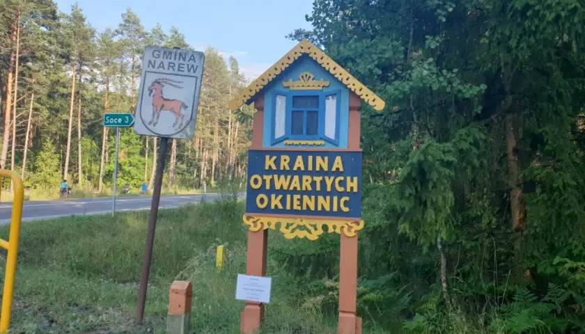 Drewniana tablica wita w Krainie Otwartych Okiennic. Obok znak Gminy Narew.