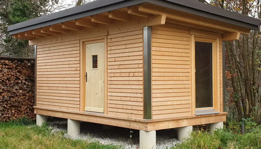 Moderne gartenhaus ideen: ein Holzhaus auf Betonpfeilern mit Pultdach, Holztür und großem Fenster. Daneben ein Holzstapel.
