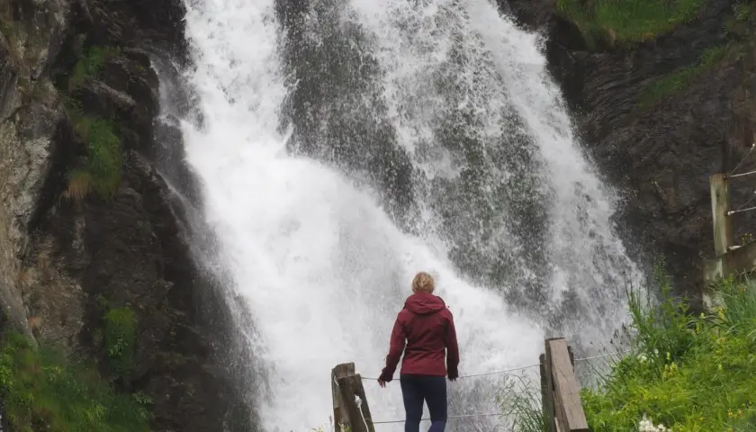Mujer contemplando una cascada en Vielha e Mijaran. El agua cae con fuerza sobre rocas oscuras, rodeada de vegetación verde.