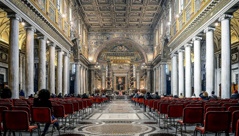 Intérieur de Sainte-Marie-Majeure, Rome. Des fidèles assis sur des chaises rouges, sous des colonnes imposantes et un plafond orné.