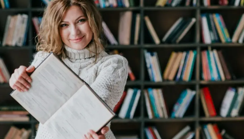 Kobieta z książką w bibliotece. Dowiedz się, jakie są zasady dystrybucji książek do bibliotek publicznych i szkolnych.