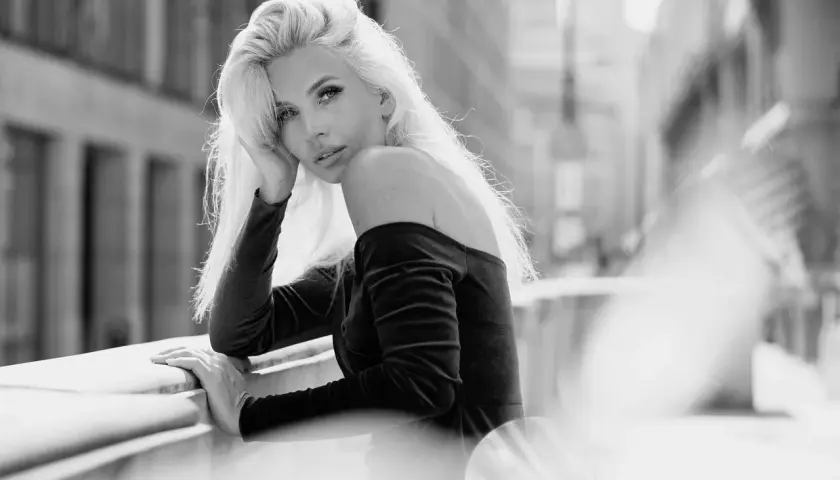 Elegancki portret w plenerze. Kobieta z blond włosami opiera się o balustradę, patrząc w obiektyw.