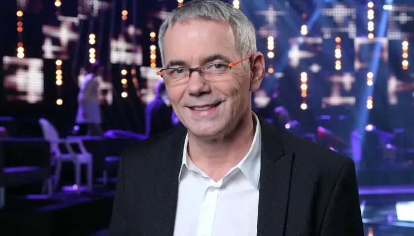 Uśmiechnięty Robert Janowski w okularach na tle sceny. Wiele osób zastanawia się, ile lat ma Robert Janowski, ale jego wiek nie jest widoczny na zdjęciu.