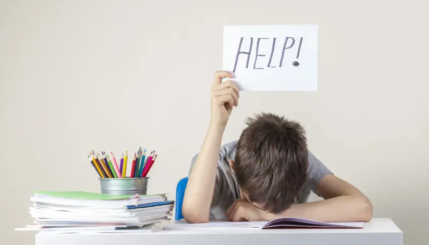Chłopiec z kartką "HELP!" nad głową, przy biurku pełnym książek i kredek. Może to być sygnał trudności, wymagający diagnozy psychologicznej.