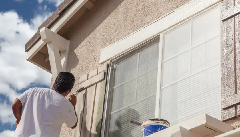 Pintor aplica pintura a persianas de ventana. ¿Cuál es la mejor pintura para fachadas? Busca durabilidad y resistencia a la intemperie.