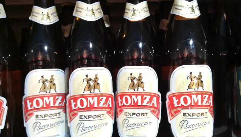 Rząd butelek piwa Łomża Export Pszeniczne. Etykiety ozdobione postaciami w strojach ludowych.