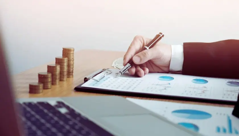 Ręka pisze na dokumentach z wykresami, obok stosy monet. Sąd może żądać wyciągu z konta w sprawach finansowych.