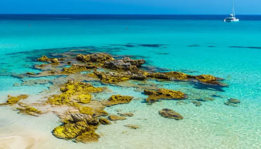 Eaux turquoise et rochers dorés sur une plage du sud de l'Italie. Un voilier blanc navigue au loin.