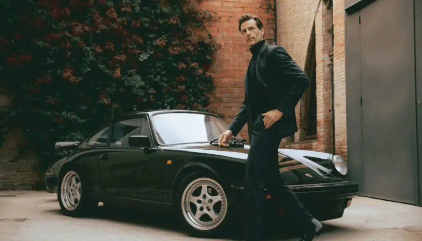 Un homme élégant en costume et bottines noires pose près d'une Porsche verte.