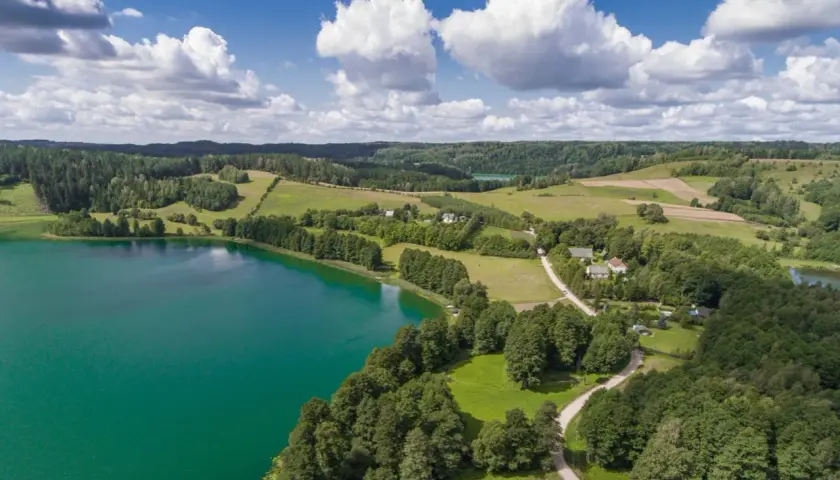 Widok z lotu ptaka na turkusowe jezioro, zielone wzgórza i lasy, z kilkoma domami.