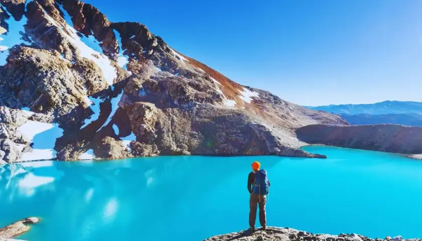 Sender solitario contempla un lago turquesa en la patagonia chilena, rodeado de montañas nevadas y un cielo azul intenso.