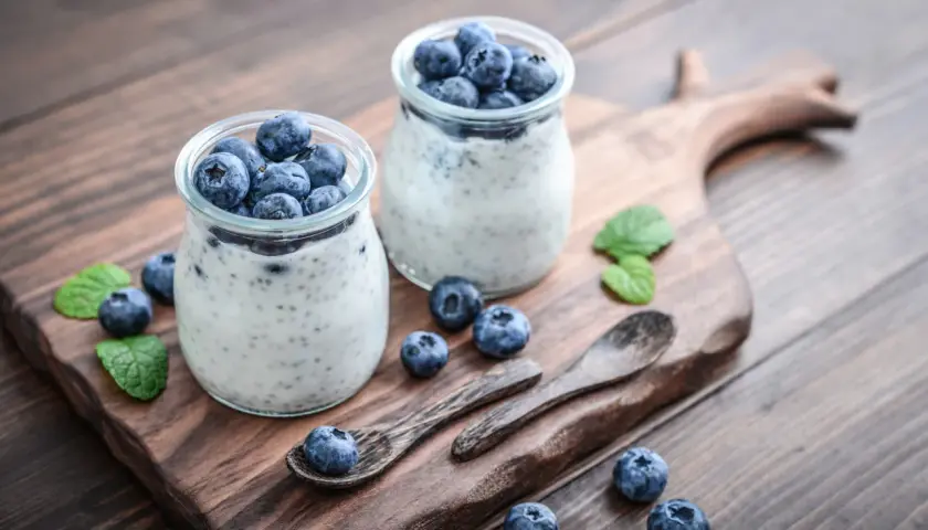 Dwa słoiczki z deserem pudding chia, ozdobione świeżymi borówkami i listkami mięty, na drewnianej desce.