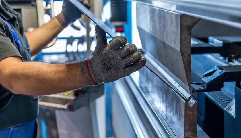 Ręce w rękawicach trzymają blachę, która jest formowana na prasie krawędziowej. To pokazuje, co to jest metal – materiał poddany obróbce.