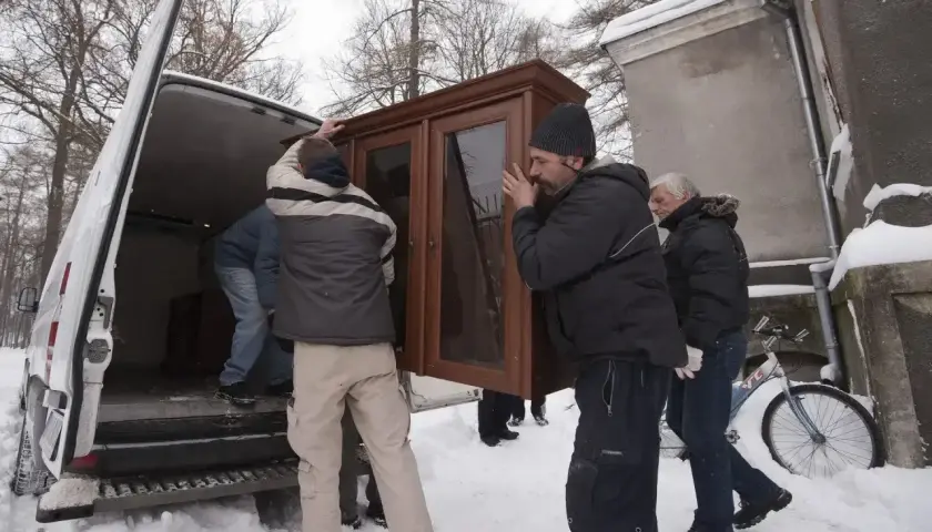 Mężczyźni wynoszą mebel z domu, przygotowując się do przeprowadzki. Widać śnieg i zimową aurę, co może sugerować trudności związane z eksmisją w okresie ochronnym.