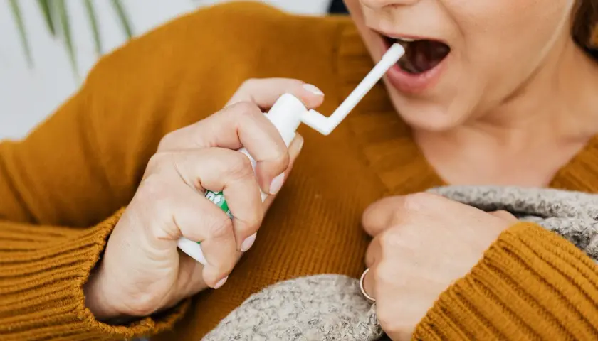 Une personne utilise un spray buccal pour soulager un mal de gorge. Elle tient le flacon blanc et dirige l'embout vers sa bouche ouverte.