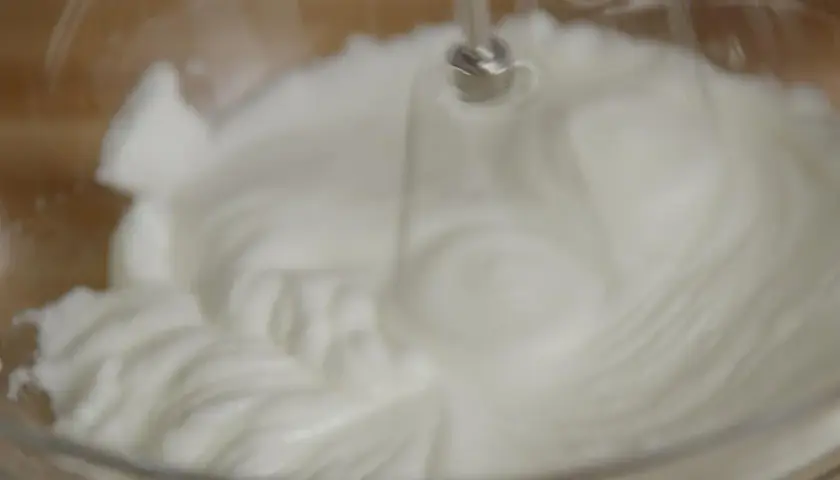 Un batteur électrique fouette un œuf blanc en neige dans un bol en verre, créant une texture aérienne et mousseuse.