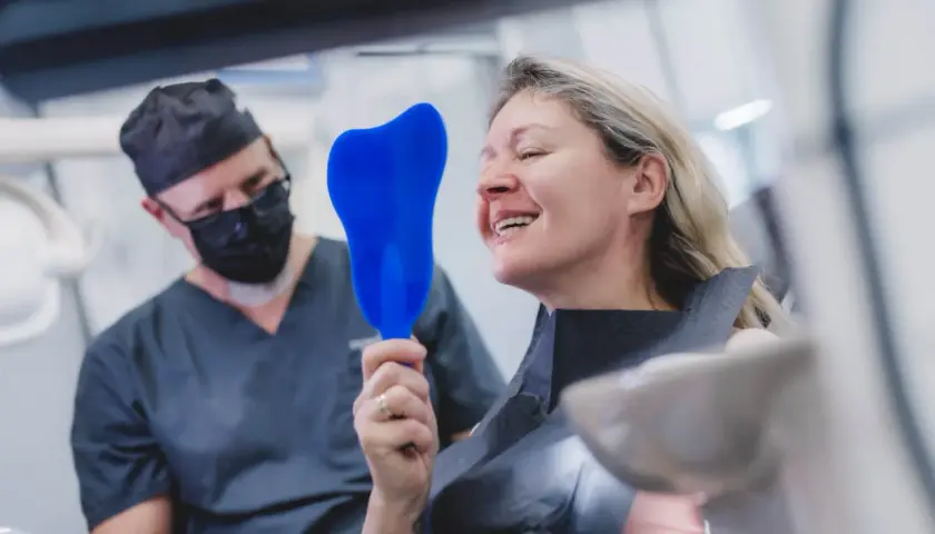 Uśmiechnięta pacjentka ogląda swoje zęby w lusterku, podczas gdy dentysta w masce obserwuje. Czy po wyrwaniu zęba można lecieć samolotem? Zapytaj specjalistę.