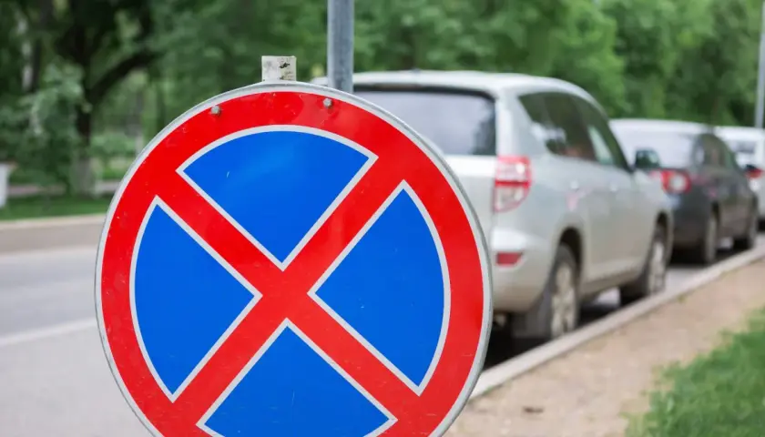 Znak zakazu zatrzymywania się b36. Samochody stoją w rzędzie przy ulicy, ale ten znak przypomina, że nie wolno tu parkować.