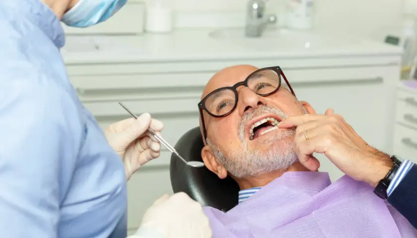 Dentysta w masce i rękawiczkach bada uśmiech starszego mężczyzny w okularach.