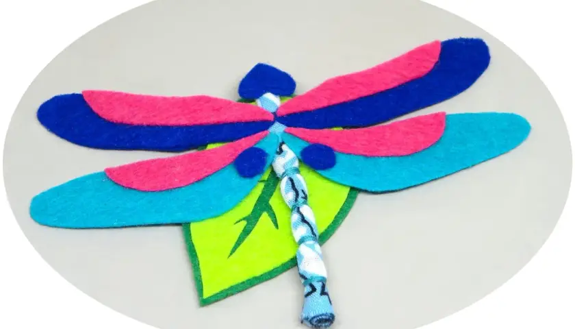 Libélula de fieltro, un proyecto de manualidades con alas azules, rosas y turquesas, posada sobre una hoja verde.