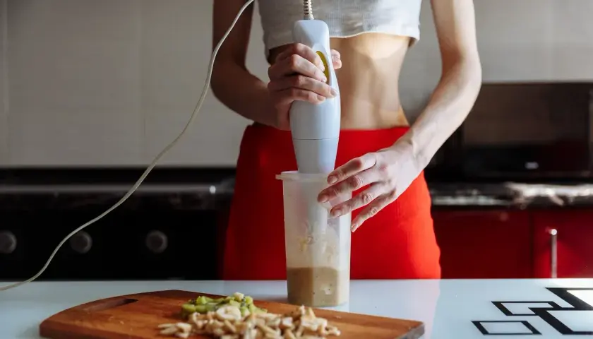Kobieta miksuje składniki na shake'a blenderem ręcznym. Na desce pokrojone owoce.