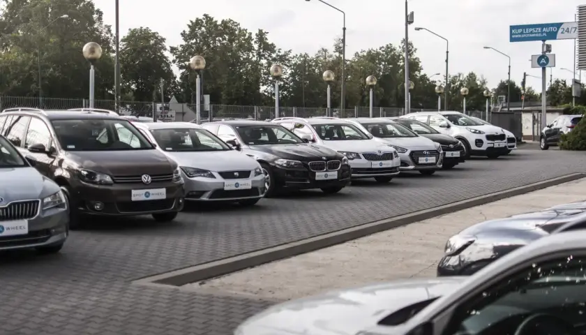 Rząd samochodów na placu, w tym brązowy Volkswagen i biały Kia Sportage. W tle drzewa i słupy oświetleniowe. To idealne miejsce, by znaleźć swój wymarzony kinto one.