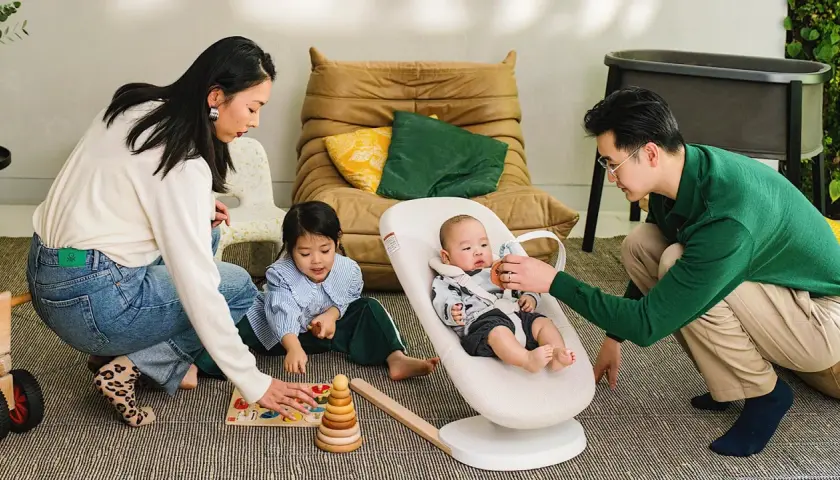 Familjen leker på golvet. Bebisen sitter i en Stokke babysitter medan pappan leker med den. Systern leker med en träleksak.