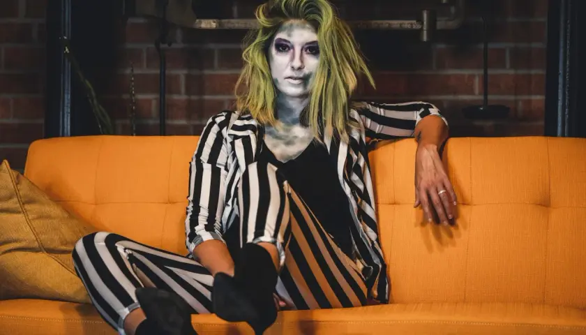 Donna con trucco spettrale e capelli verdi, indossa un vestito da strega fai da te a righe bianche e nere, seduta su un divano arancione.