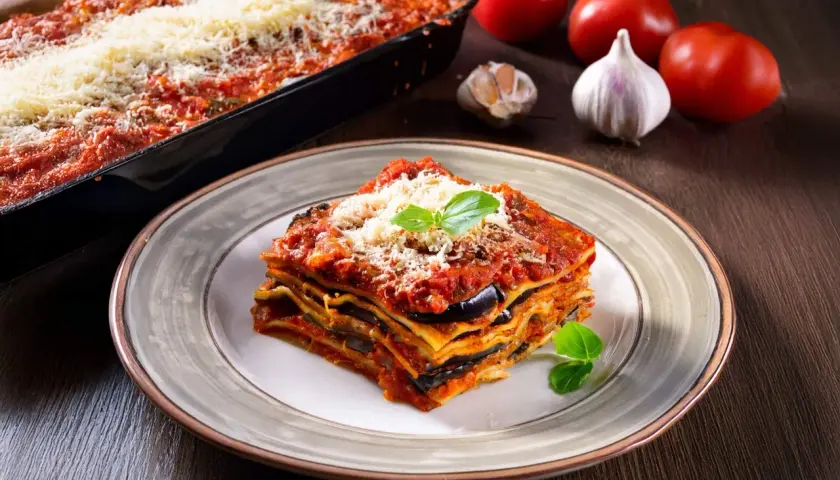 Una fetta di lasagne vegetariane, con melanzane e pomodoro, guarnita con basilico e parmigiano. Sullo sfondo, ingredienti freschi.