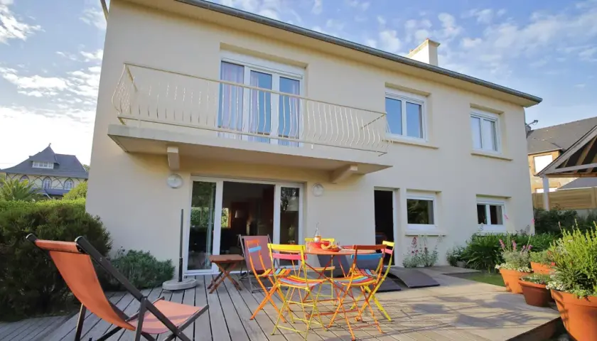 Maison divisée en 2 logements avec terrasse en bois, mobilier coloré et transat. Jardin verdoyant et ciel bleu.